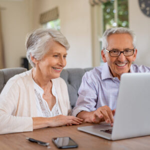 Happy smiling retired couple using laptop at home. Cheerful elderly man and old woman using computer while sitting at table. Smiling pensioner showing woman notebook at home.